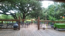 Wooden deck with multiple empty metal tables and chairs, covered by large trees. A green tablecloth is in the right corner.
