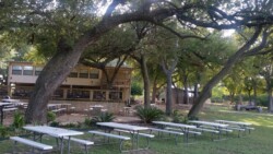 Outdoor area with white picnic tables under large trees, in front of a two-story rustic building with a shaded porch.